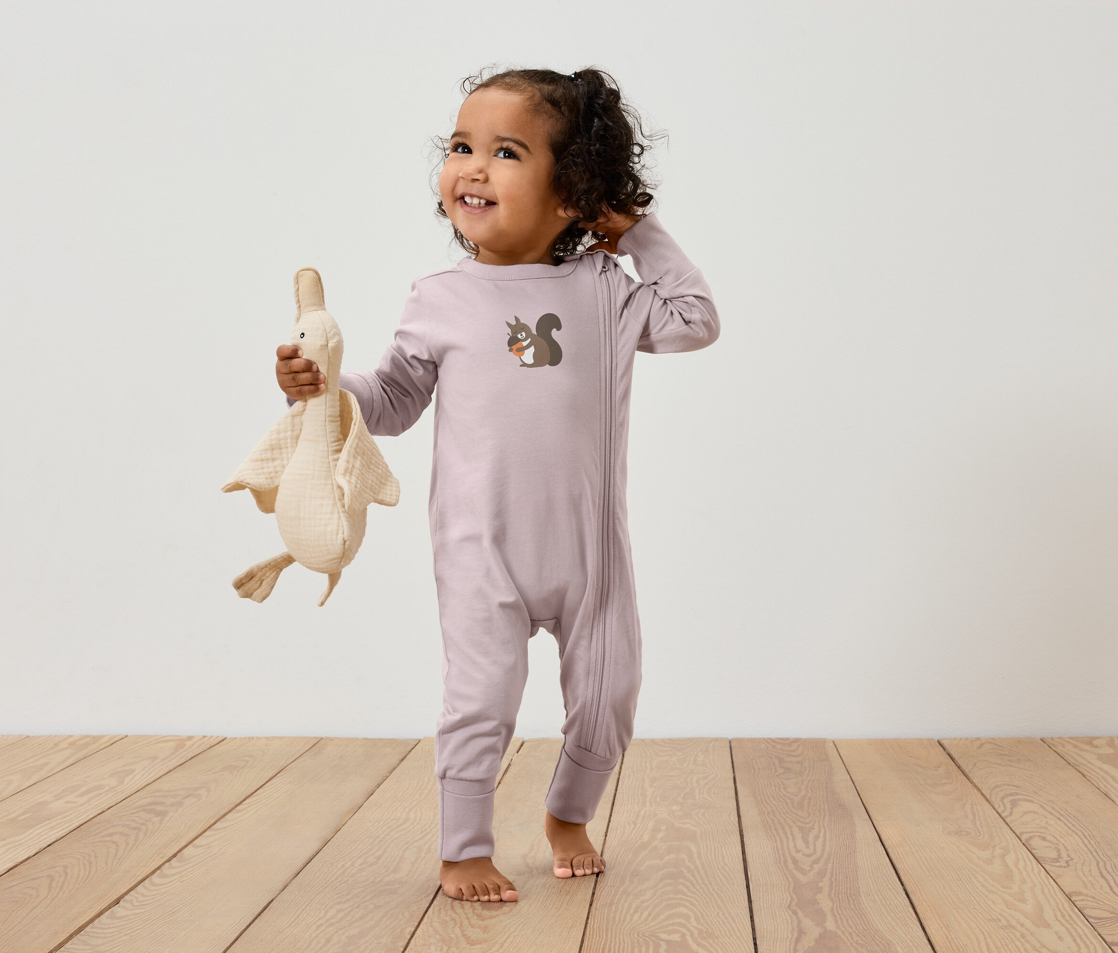 Egy rózsaszín, cipzáros, mókus mintásOverallban lévő gyermek egy liba plüssjátékot tart.