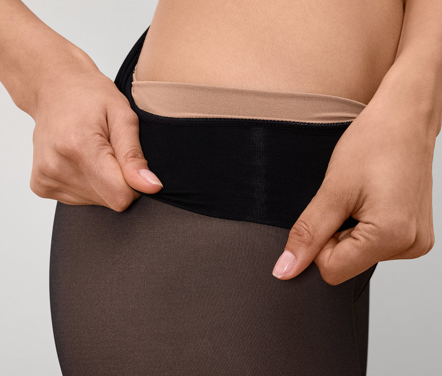 Egy nő közeli képe, aki fekete harisnyanadrágot húz fel, testszínű derékrésszel.