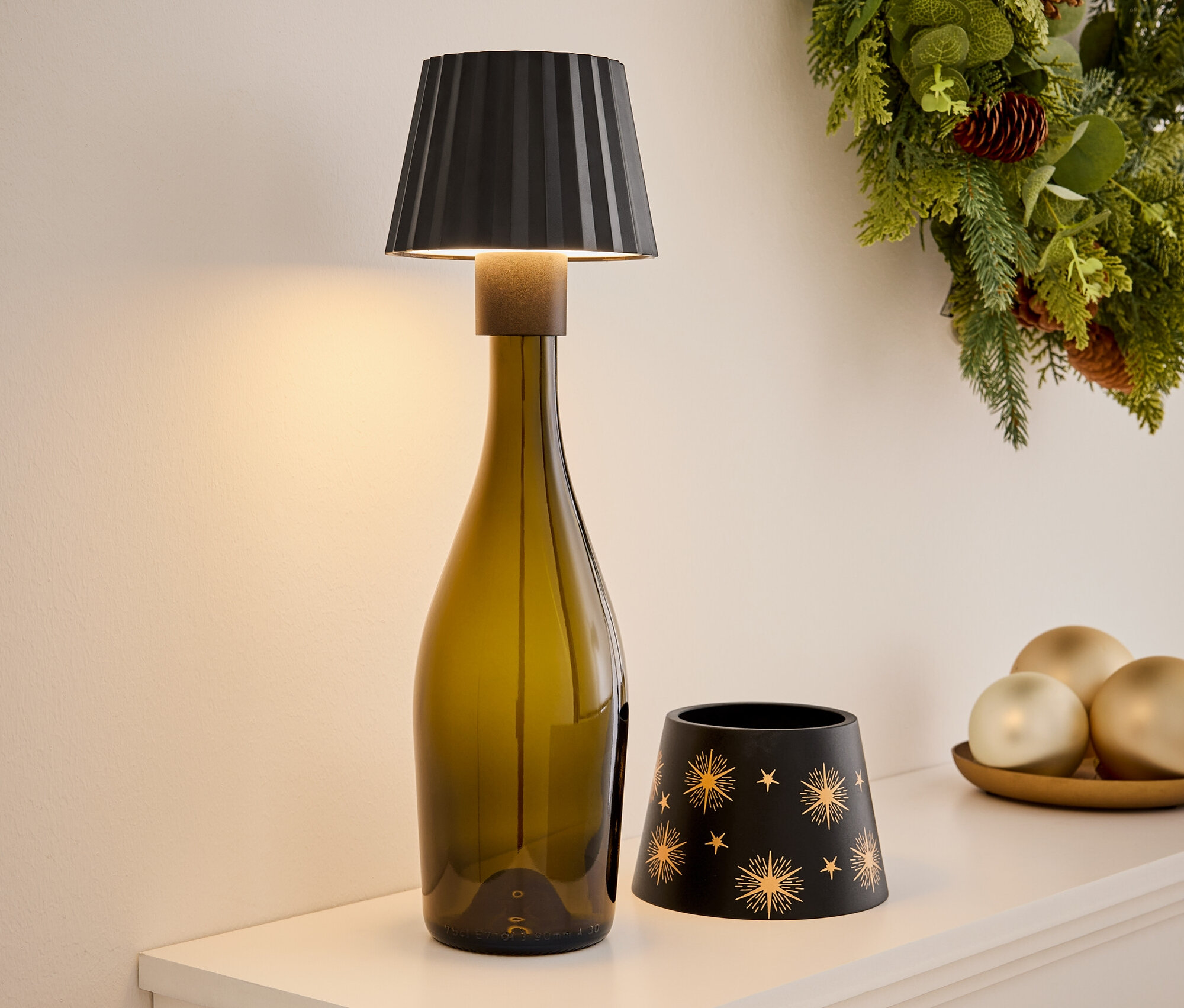 Egy fehér polcon egy olívazöld üvegből készült lámpa, egy fekete, arany csillagokkal díszített gyertyatartó és díszek egy tálcán. A háttérben karácsonyi koszorú látható.