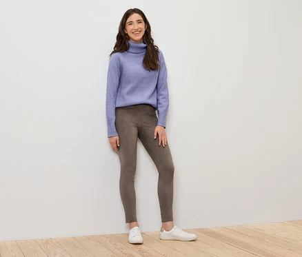 Egy nő áll, lila garbót, barna leggingset és fehér tornacipőt visel.