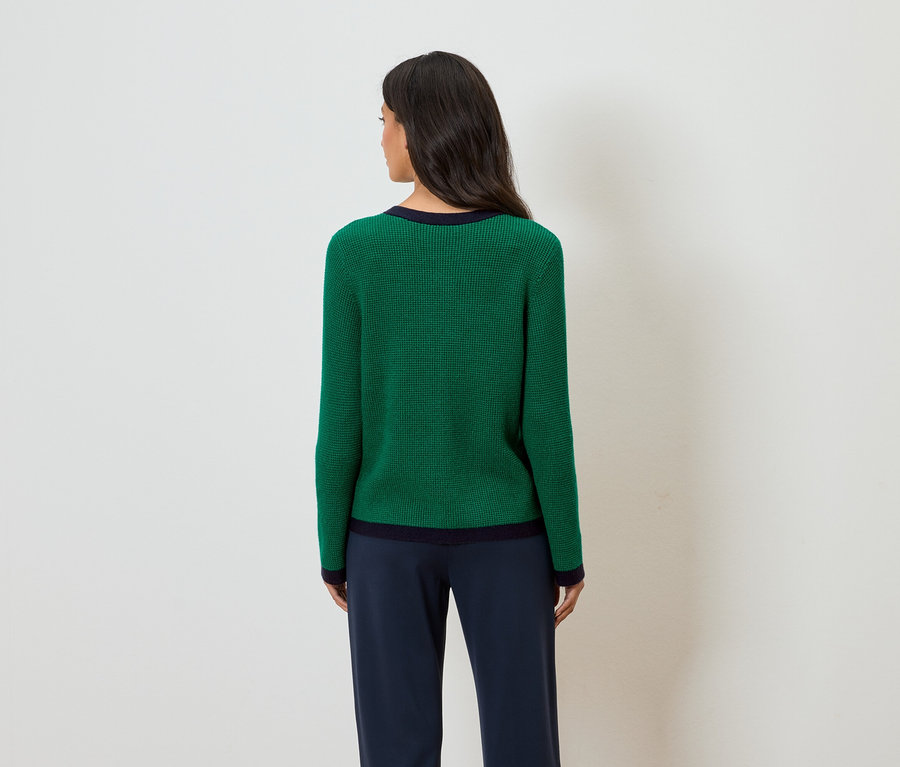 Hátulnézet egy hosszú, sötét hajú nőről, aki tengerészkék szegélyű zöld pulóvert és tengerészkék nadrágot visel.