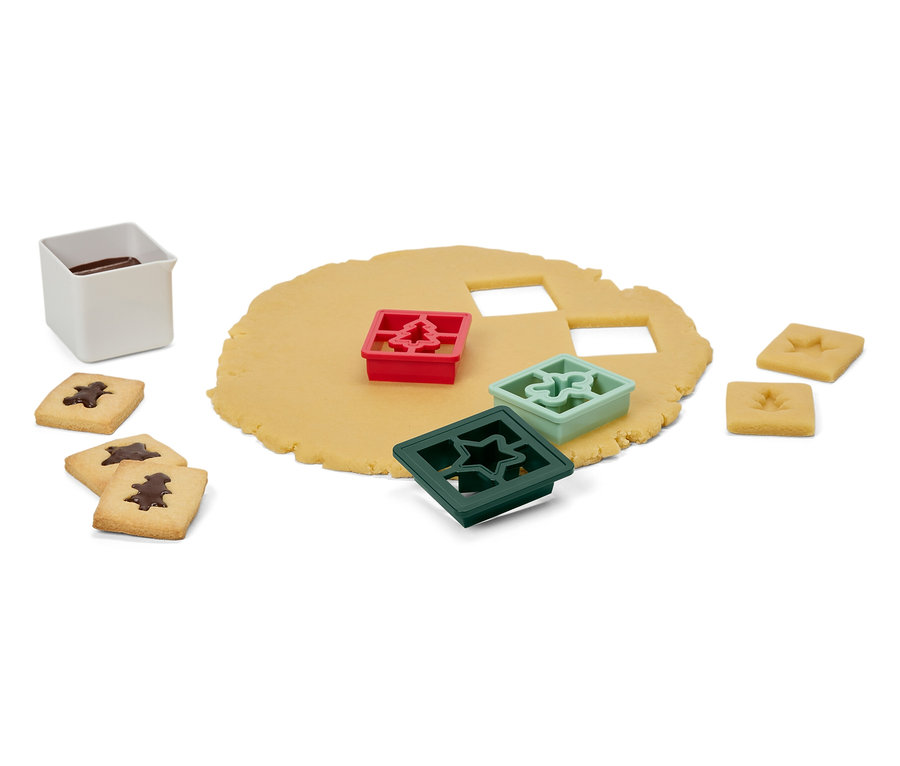 Tészta fa, csillag és dísz alakú kiszúrókkal, kész sütik és egy fehér edény mázzal.