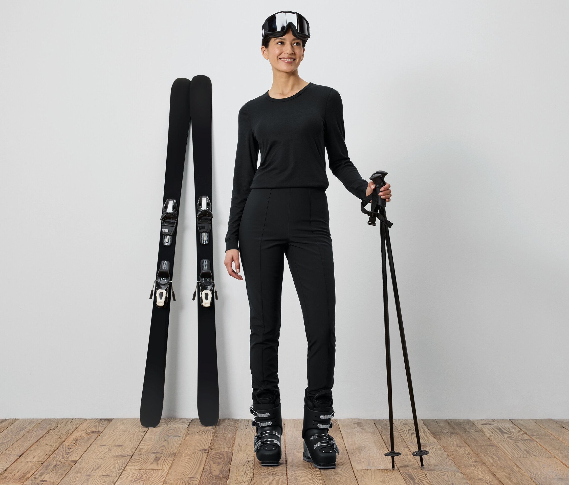 Fekete síruhát viselő nő áll a sílécek és botok mellett.