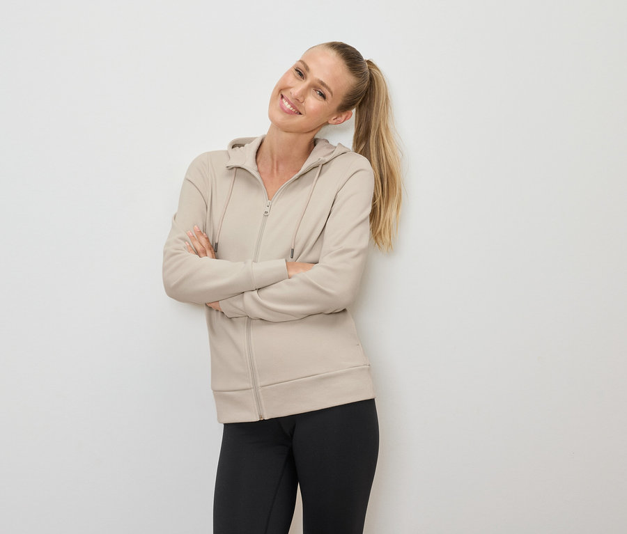 Mosolygó szőke, copfos nő keresztbe tett karokkal pózol, bézs színű, cipzáras kapucnis pulóvert és fekete leggingst visel.