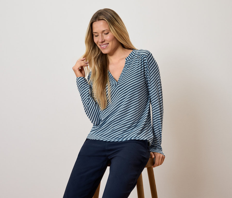 Nő ül egy széken, kék-fehér csíkos, hosszú ujjú felsőt és sötétkék nadrágot visel.