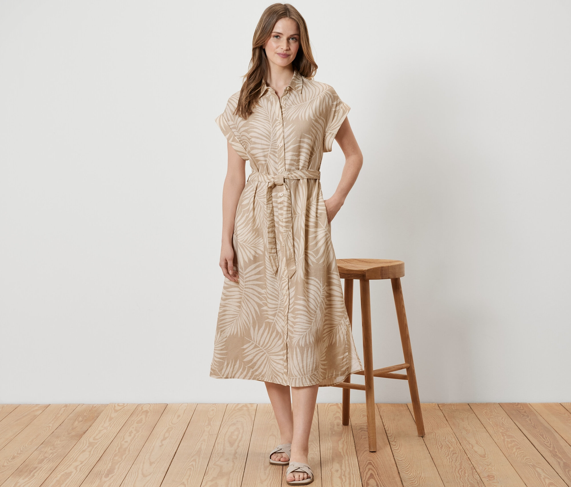 Egy nő egy fa szék mellett áll, és nyomott mintás lenvászon ruhát visel.