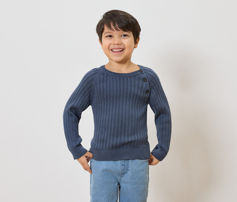 Egy mosolygó, barna hajú fiú gombos, tengerészkék, bordázott pulóvert és világoskék farmert visel.