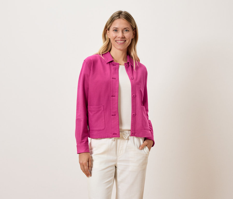 Nő visel rózsaszín kabátot egy fehér felső fölött és krémszínű nadrágot.