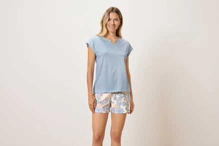 Eine Frau steht und trägt ein blaues T-Shirt und gemusterte Shorts.