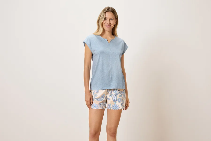 Eine Frau steht und trägt ein blaues T-Shirt und gemusterte Shorts.