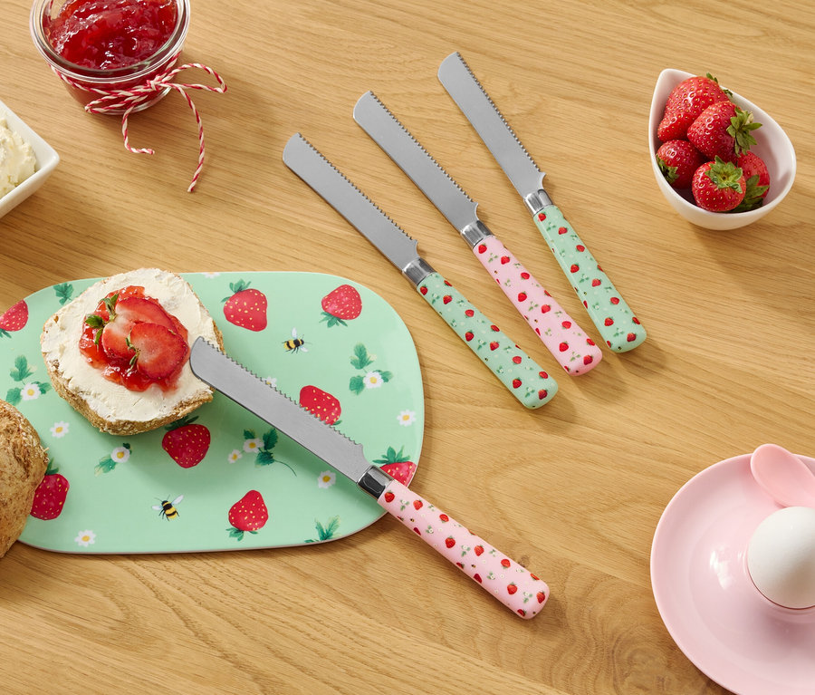 Egy fa asztalon egy zsemle, egy eper mintás tányér, krémsajt, lekvár, eper és négy eper mintás kés található.