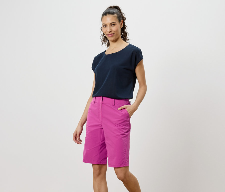 Nő sötét pólóban és rózsaszín rövidnadrágban áll fehér háttér előtt.