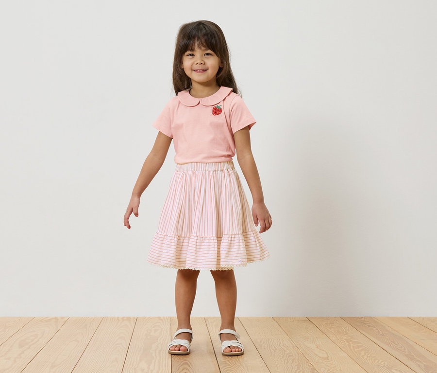 Egy kislány áll egy fehér fal előtt, rózsaszín, epres pólót és csíkos szoknyát visel.