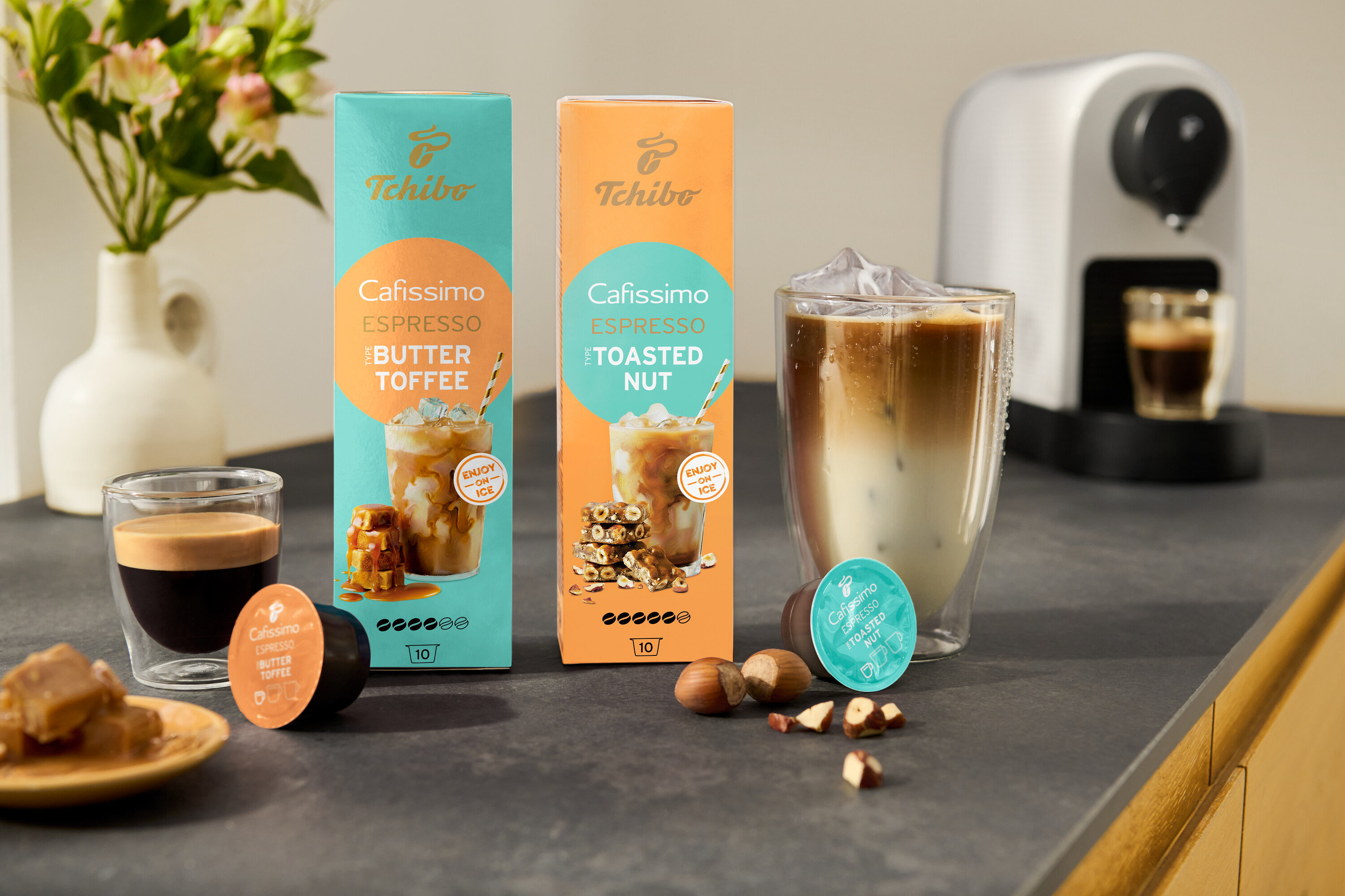 A konyhapulton két csomag Cafissimo Flavoured Espresso áll, egy türkiz színű Buttertoffee, egy narancssárga Toasted Nut ízesítésű, mellette egy fehér Cafissimo pure+ gép.