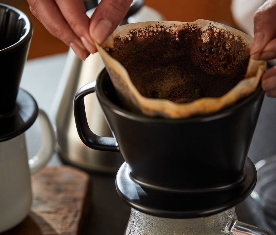 Egy nő papír szűrőt helyez egy fekete, 2-es méretű kávéfilter kézi forrázáshoz. Alatta egy kávéskanna, üveg.