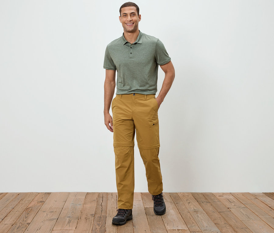 Egy férfi áll egy fehér fal előtt, zöld pólóinget és khaki nadrágot visel.