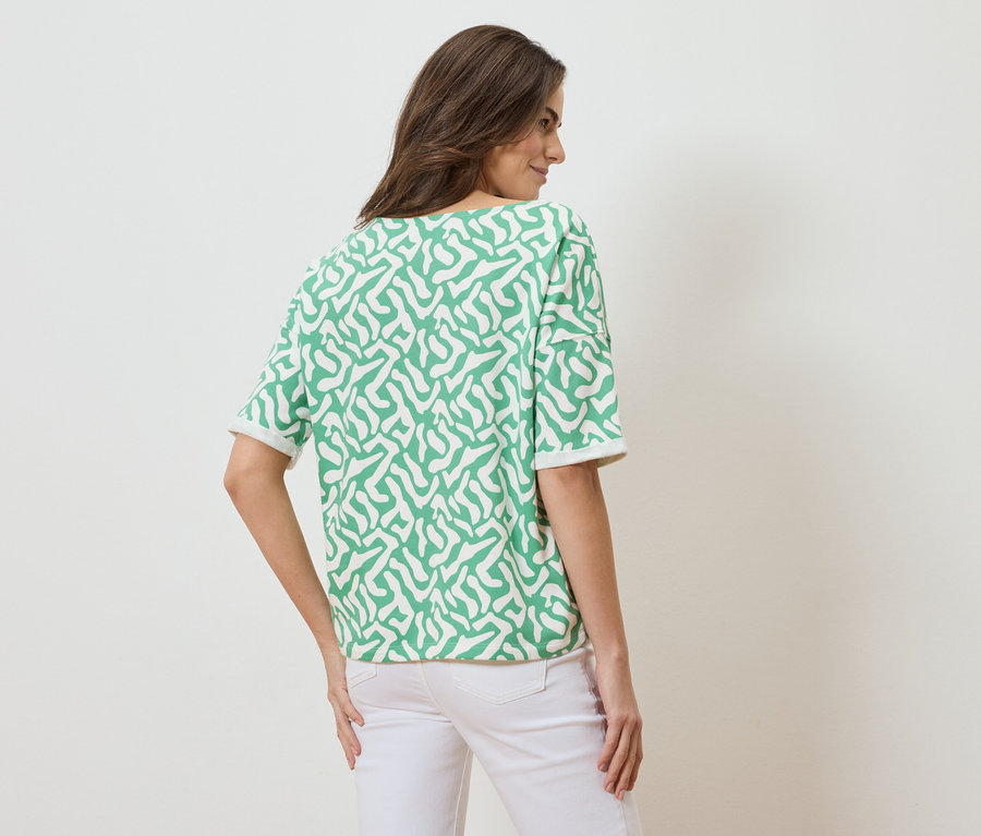 Hátulnézet egy barna hajú nőről, aki zöld-fehér mintás, rövid ujjú felsőt és fehér farmert visel.