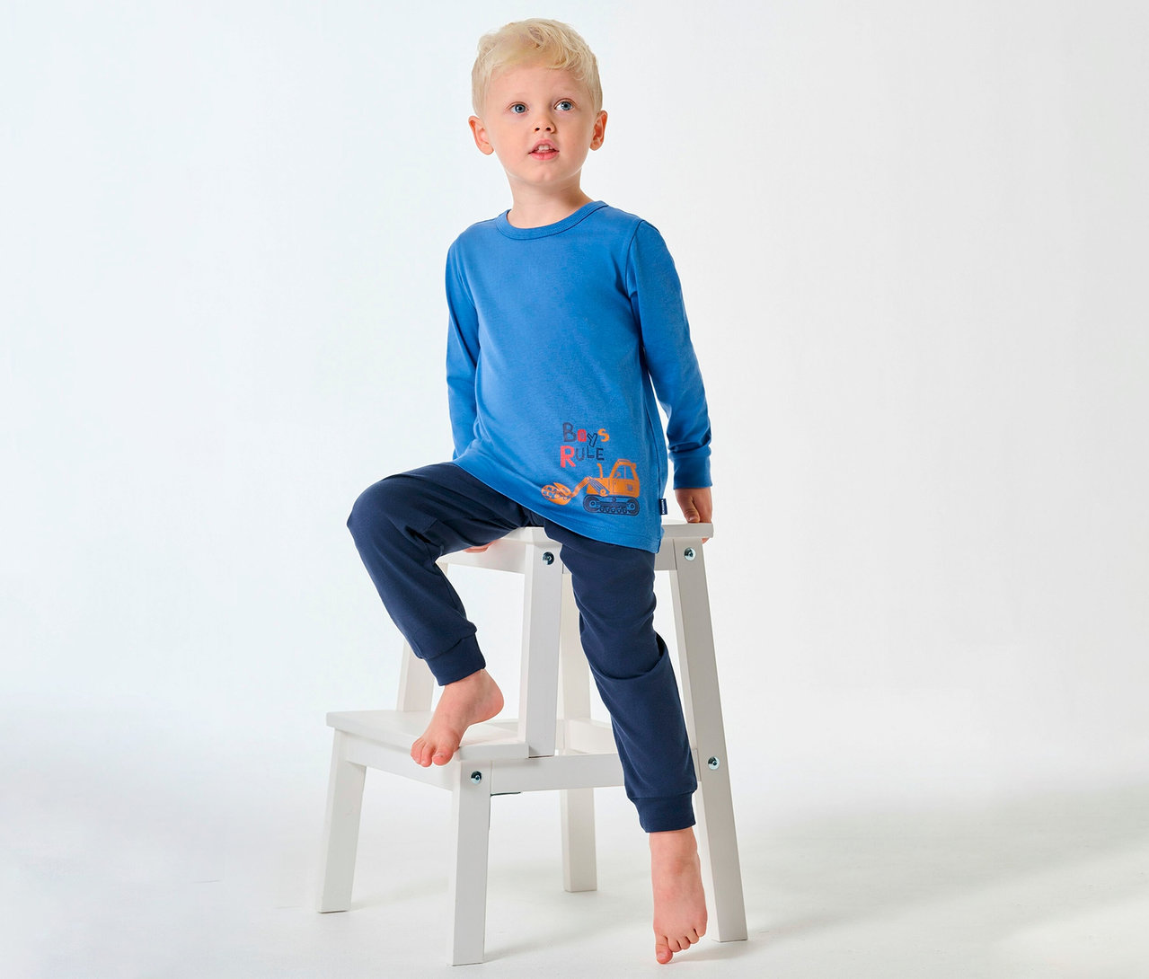 Szőke fiú SCHIESSER hosszú gyerekpizsamában ül mezítláb a sámlin.