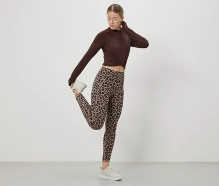 Nő nyújtja a lábát barna felsőben, leopárdmintás leggingsben és fehér tornacipőben, fehér háttér előtt.
