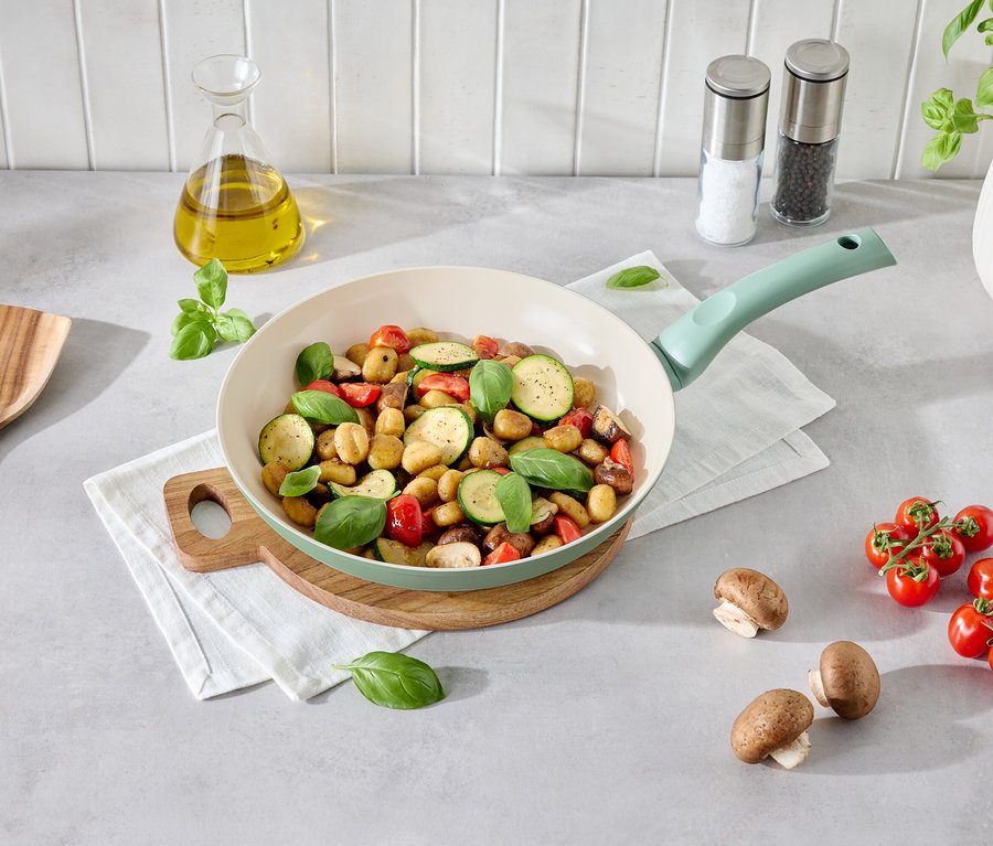 Zöldséges gnocchi egy világoszöld serpenyőben egy fa tányéron. Mellette gomba, paradicsom, fakanál és egy üveg olaj.