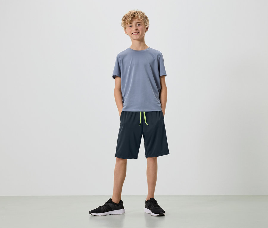 Egy szőke fürtös fiatal férfi áll fehér háttér előtt szürke pólóban, sötétkék rövidnadrágban és fekete tornacipőben.