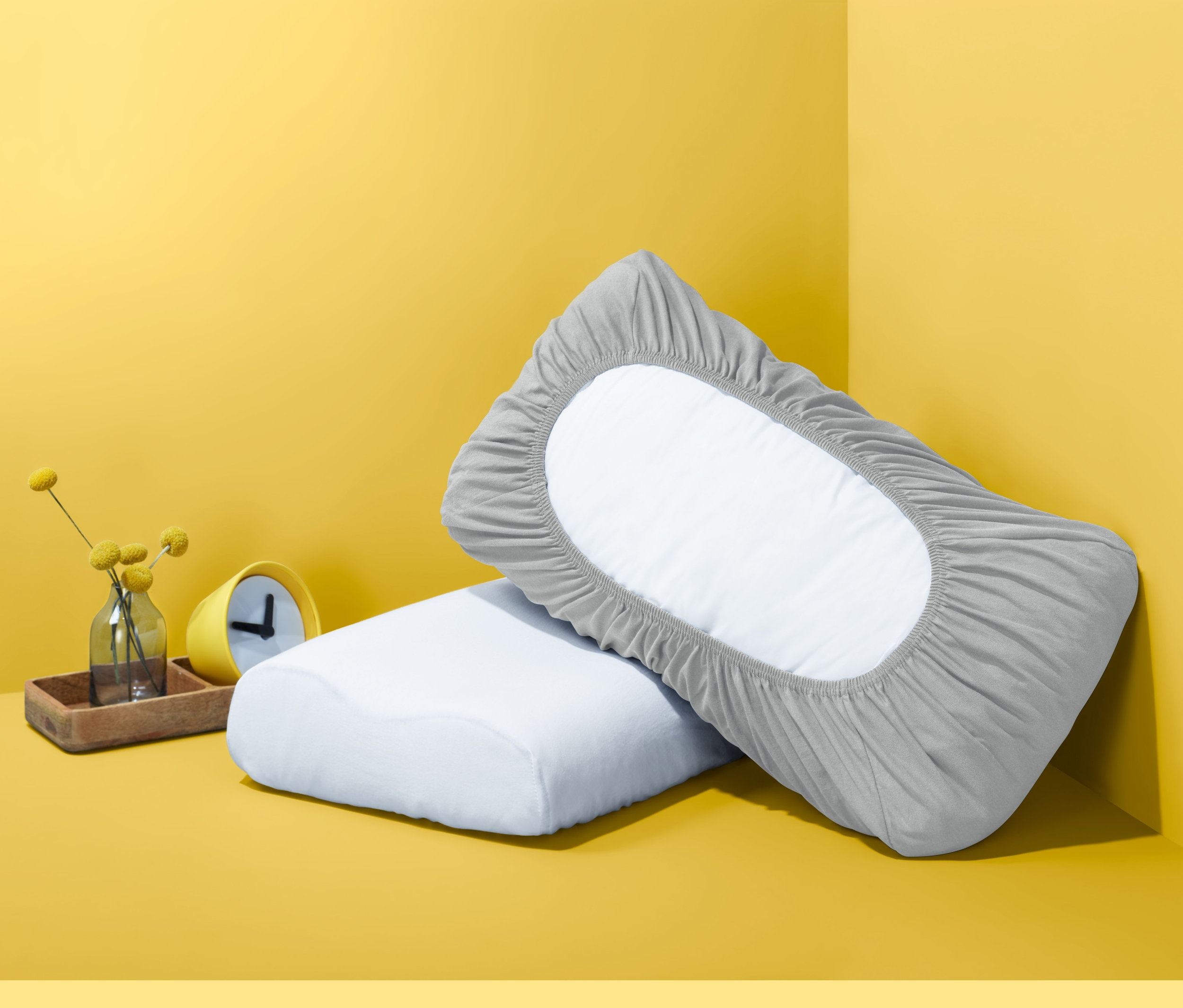 Két párna sárga háttér előtt. Az egyik párnán szürke gumis jersey párnahuzat van. 2 gumis párnahuzat szettben.