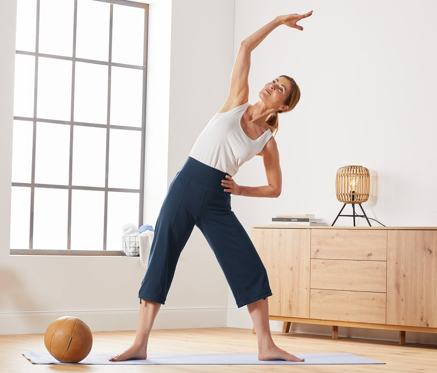 Nő nyújtó gyakorlatokat végez jógaszőnyegen, sötétkék színű, 3/4-es sportnadrágot visel.