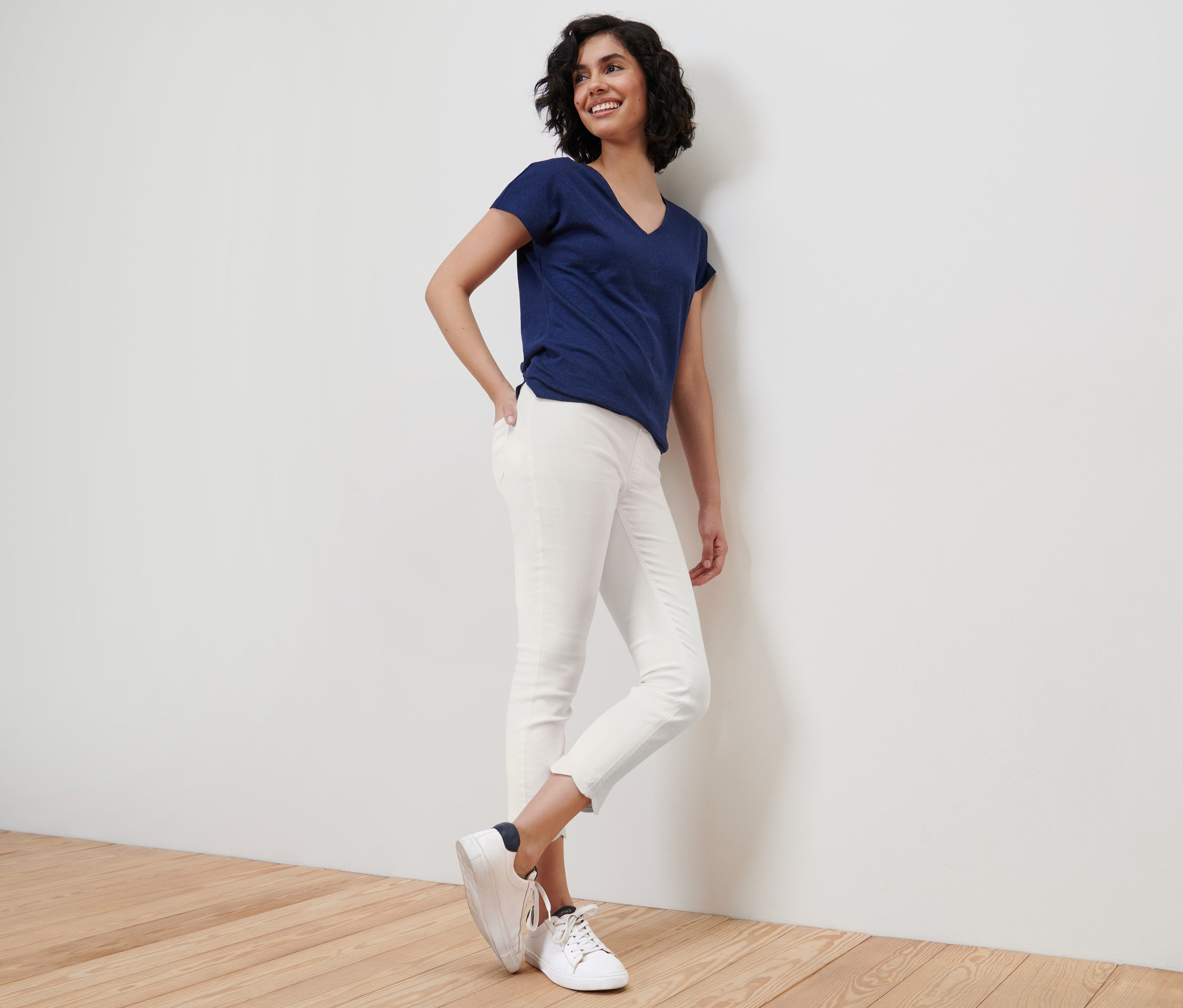 Mosolygó nő a falnak dőlve, kék pólót és fehér, 7/8-os, lyukhímzéses treggingst visel.