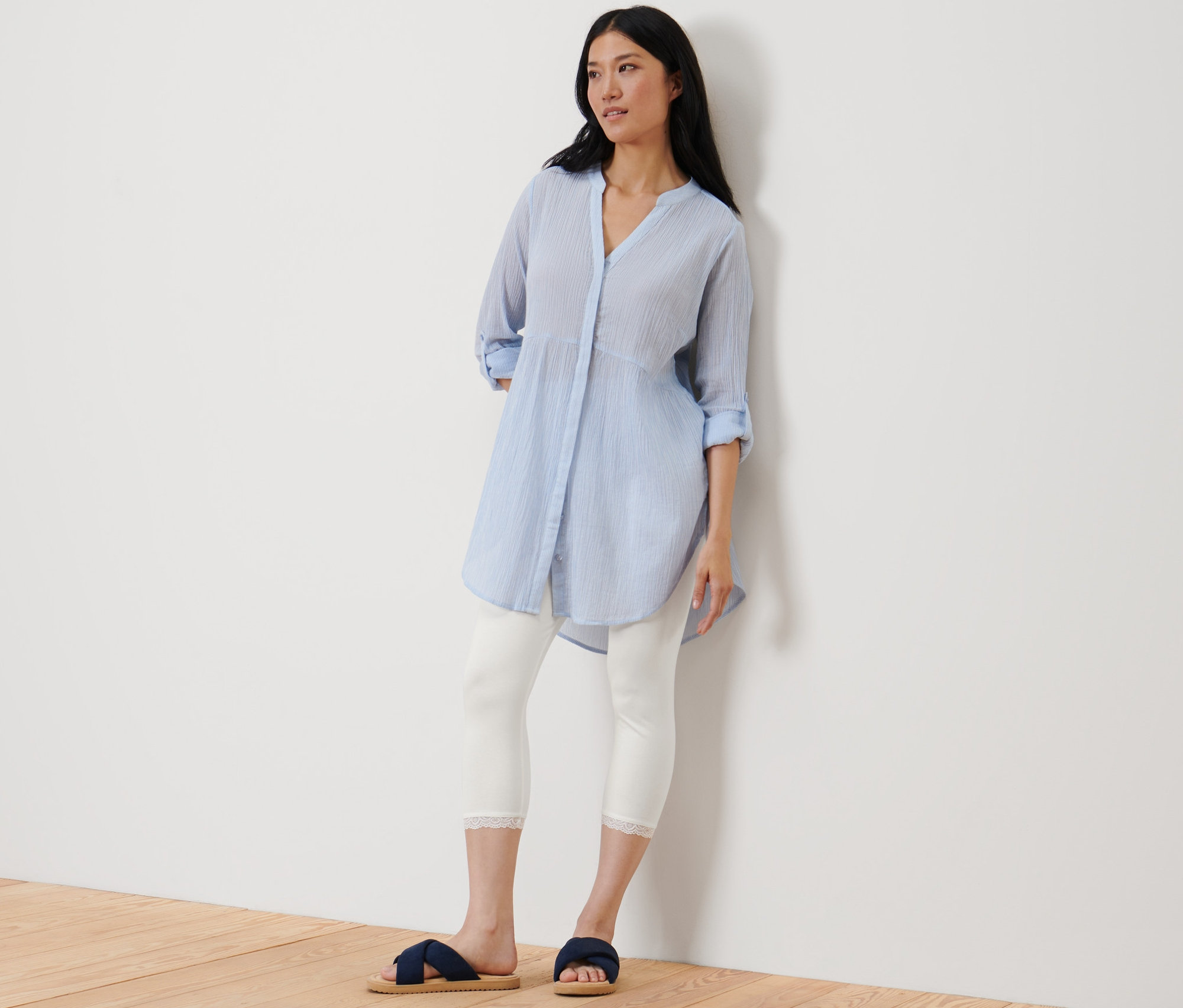 Egy nő a falnak dőlve áll, és egy fehér, csipkés szegélyű 3/4-es leggingset visel.