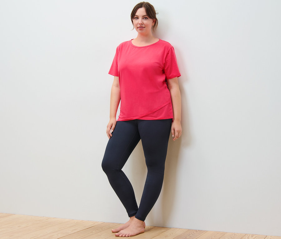 Egy nő a falnak támaszkodik, rózsaszín sportfelsőt és tengerészkék sportnadrágot visel.