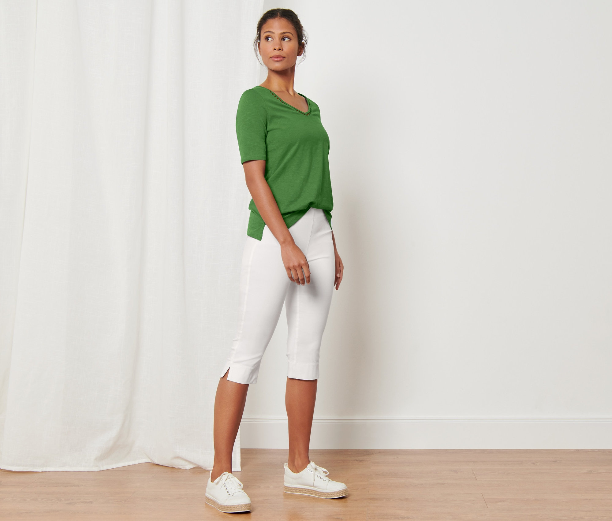Egy nő áll egy szobában, zöld pólót és fehér 3/4-es sztreccsnadrágot visel.