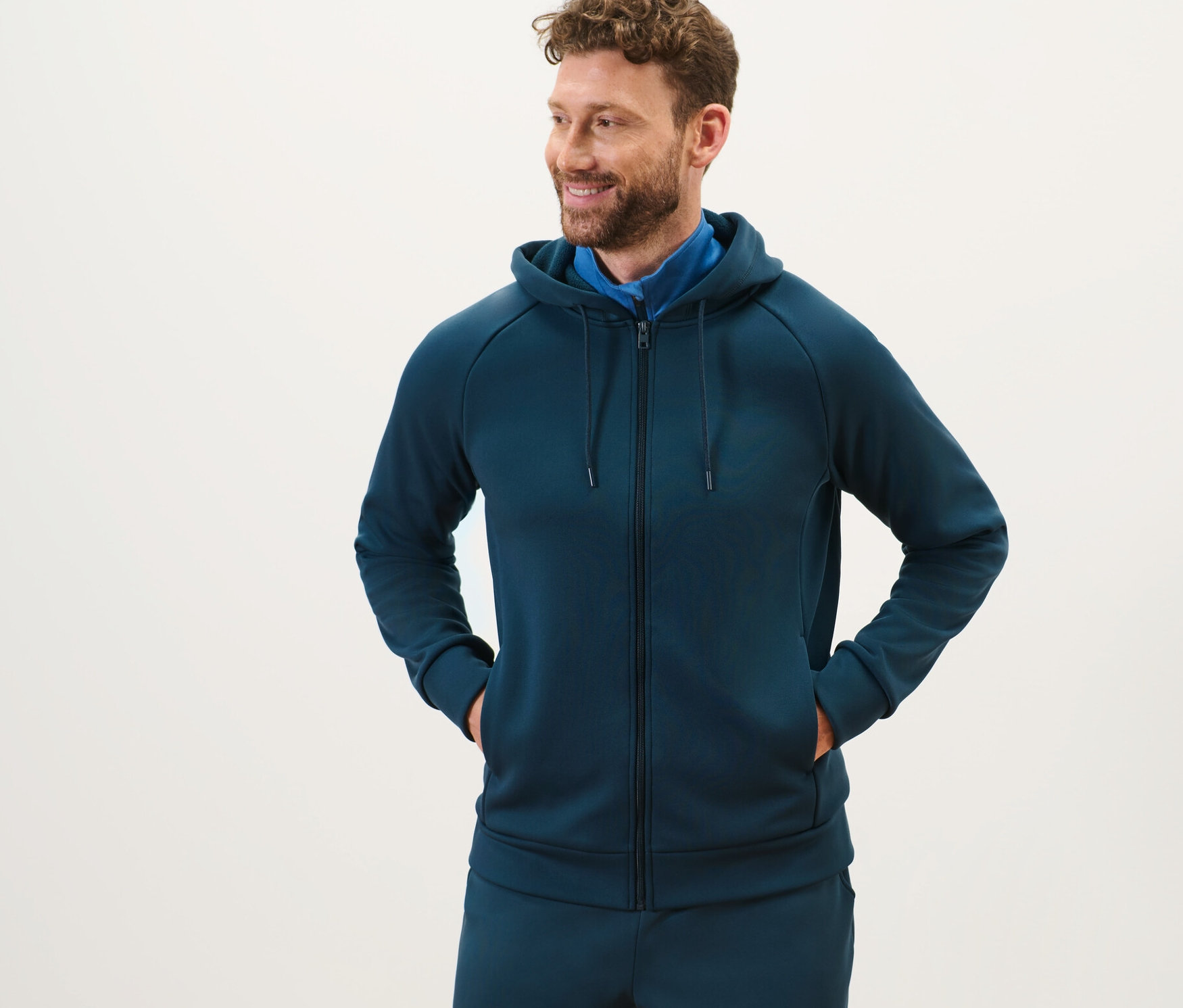Szakállas férfi áll és sötétkék férfi funkcionális kapucnis felsőt visel.