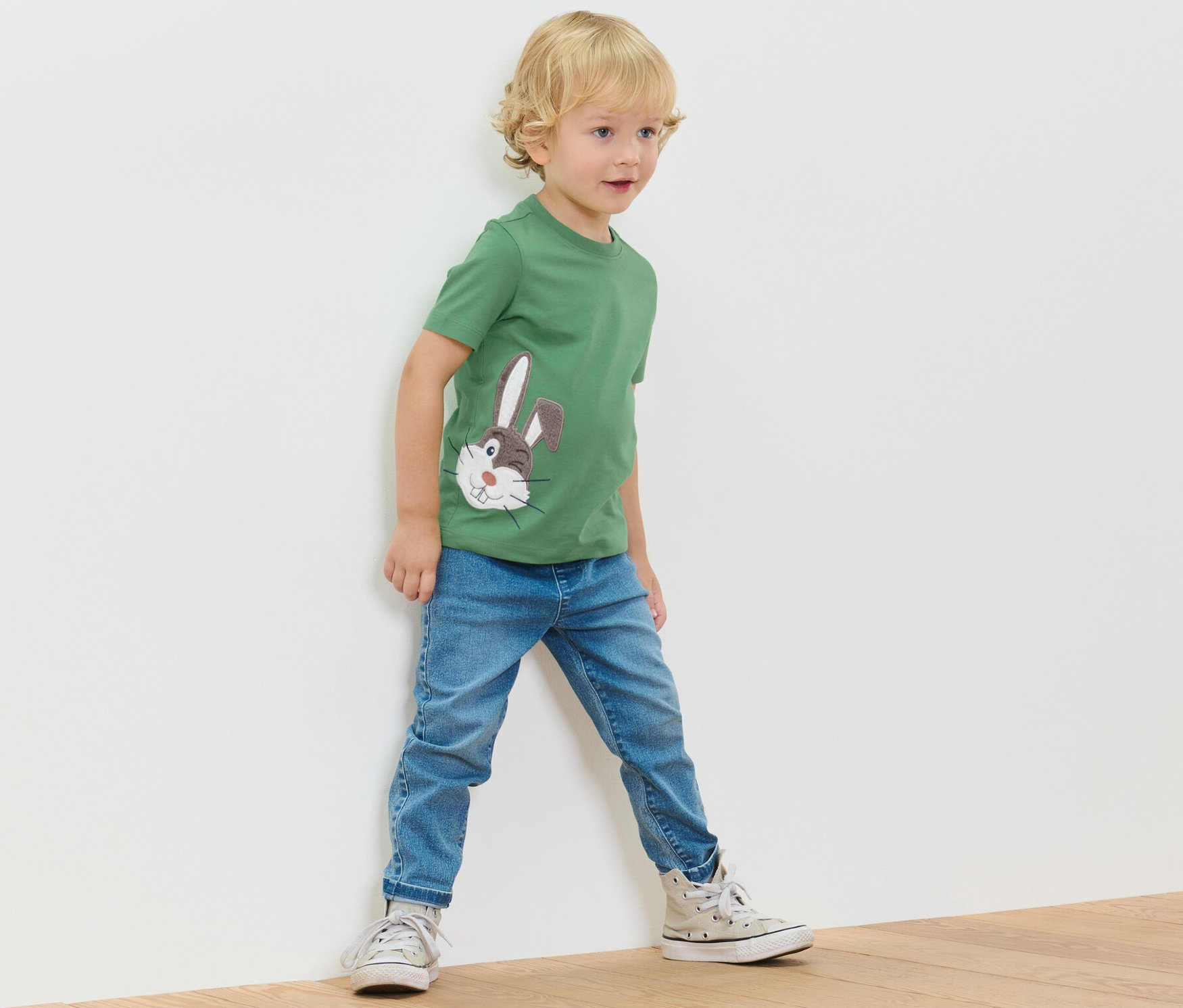 Egy kisfiú áll a fal mellett, zöld pólót és farmert visel.