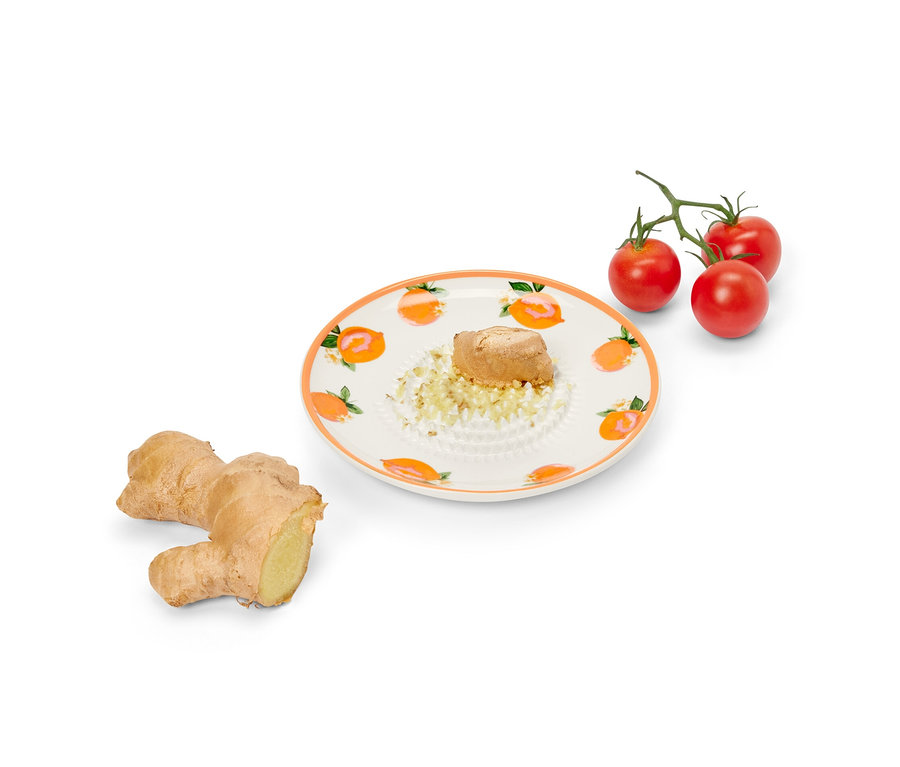 Gyömbér egy porcelán reszelőtányéron reszelve, mellette egy egész gyömbér és koktélparadicsomok.