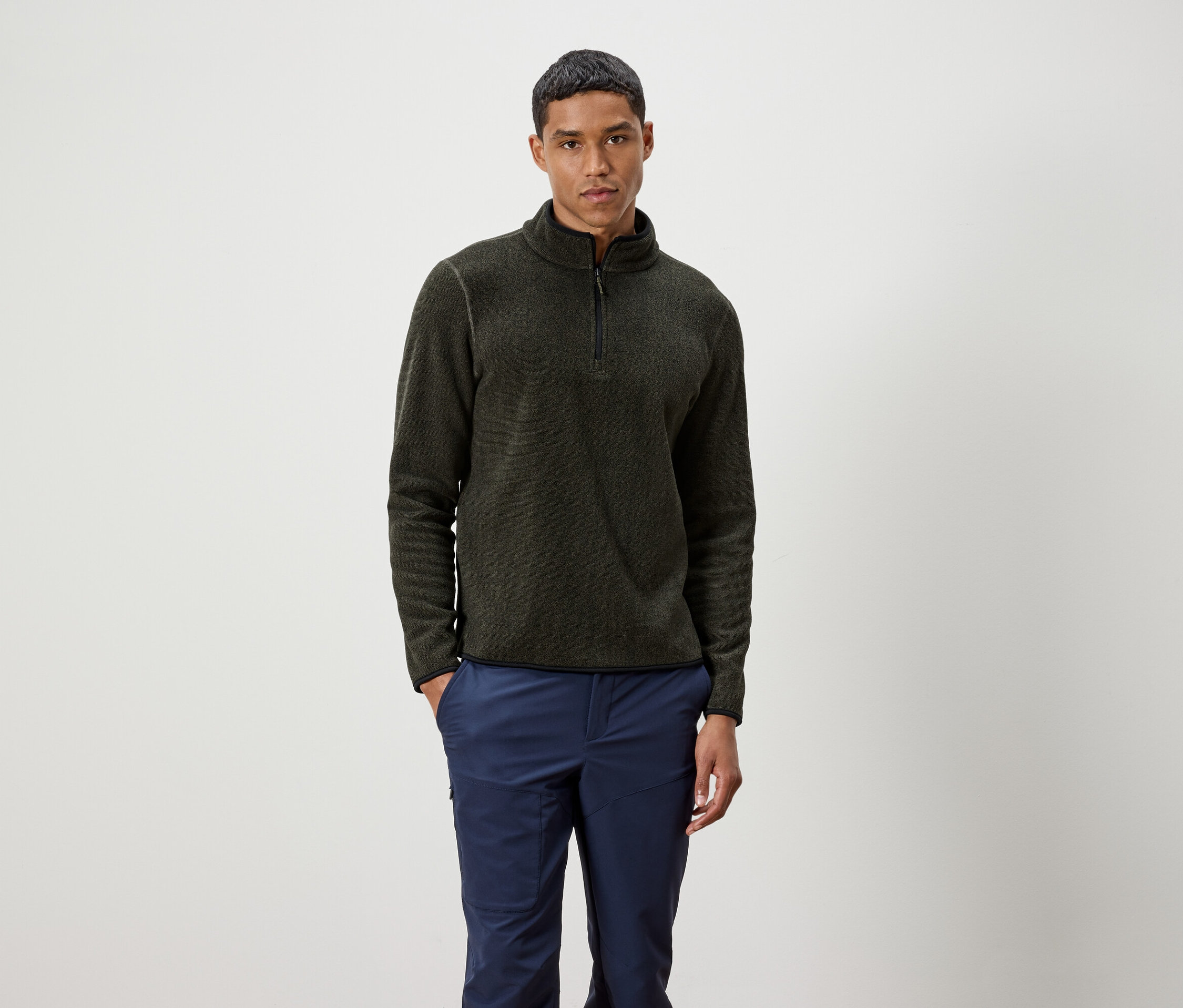 Férfi áll, olíva színű, cipzáras pulóvert és kék nadrágot visel.
