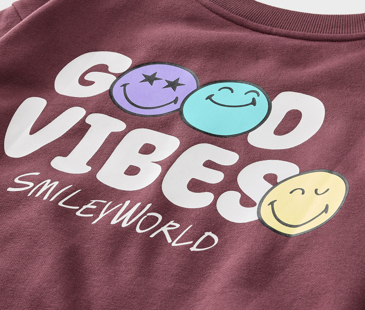 Egy bordó SMILEYWORLD® gyerek szabadidőfelsőm a belseje bolyhozott részlete, GOOD VIBES felirattal és három mosolygós arccal.