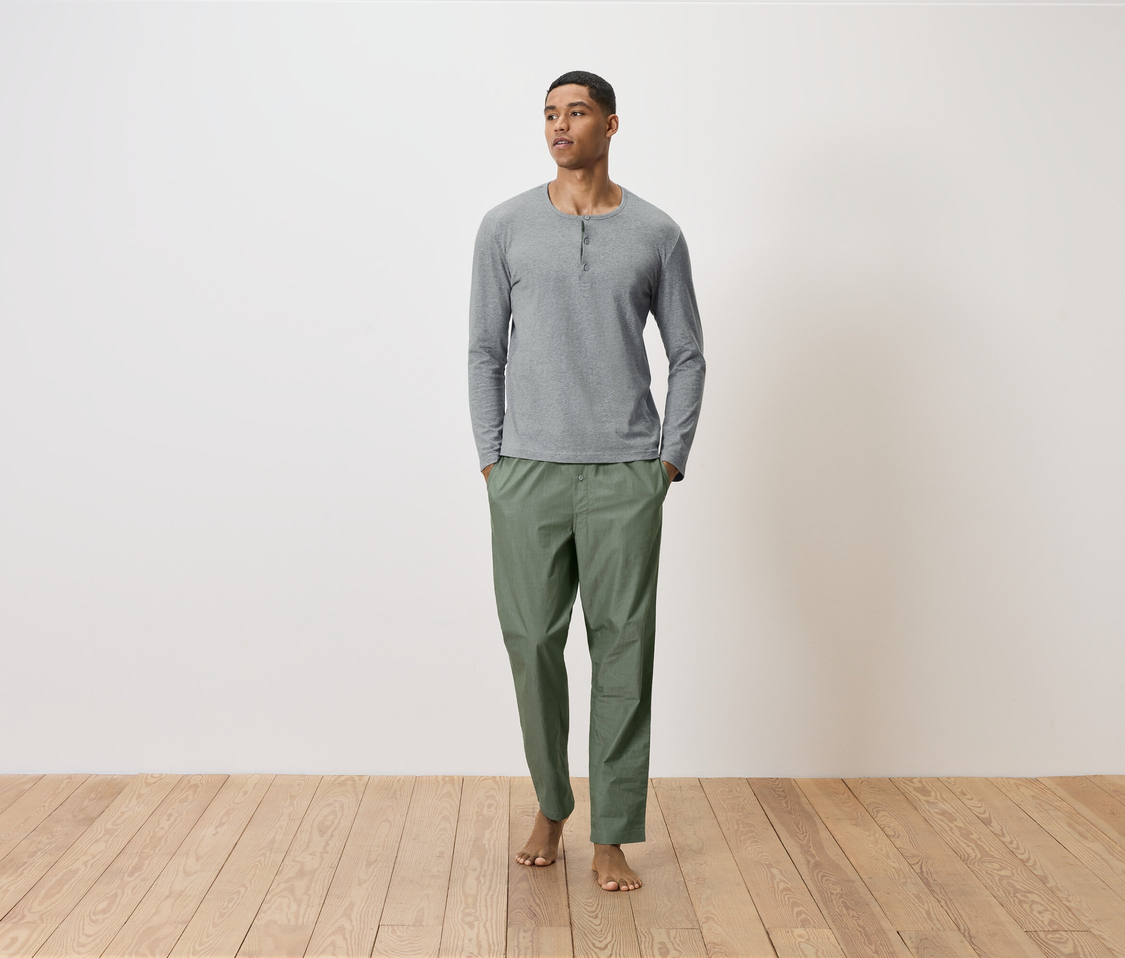 Egy férfi áll, zsebre tett kézzel, zöld nadrágban és szürke, hosszú ujjú ingben.