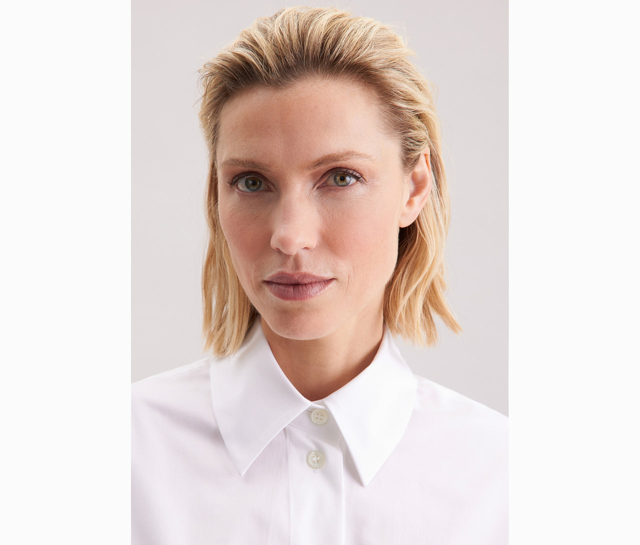 Portré egy szőke, hullámos hajú nőről, aki fehér Seidensticker túlméretes hosszú ingblúzt visel.