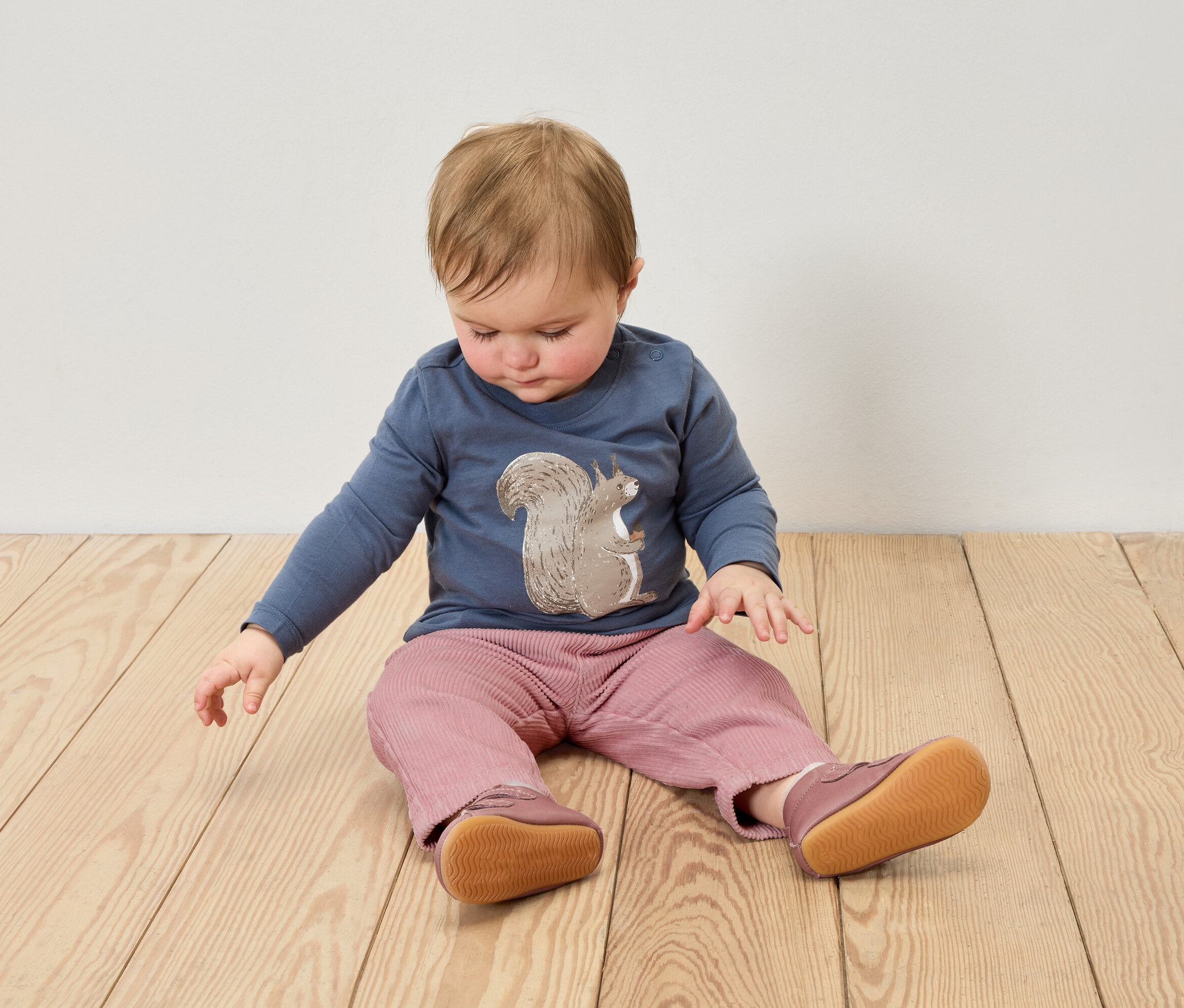 Egy kisgyermek ül a fapadlón, kék mókus mintás pólót, rózsaszín nadrágot és bőr babacipőt visel.