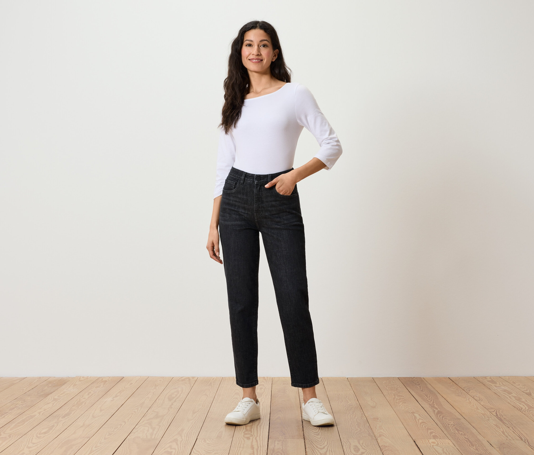Nő pózol fekete farmerban, háromnegyedes ujjú fehér felsőben és fehér tornacipőben.