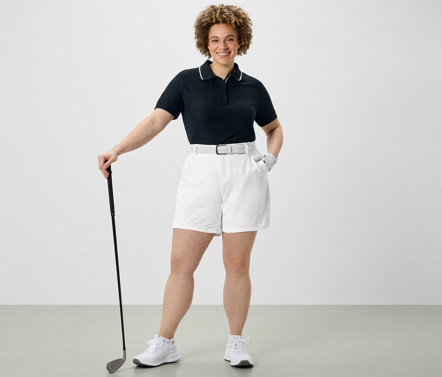 Nő golflabdát tart, sötétkék funkcionális pólóinget és fehér funkcionális softshell rövidnadrágot visel.