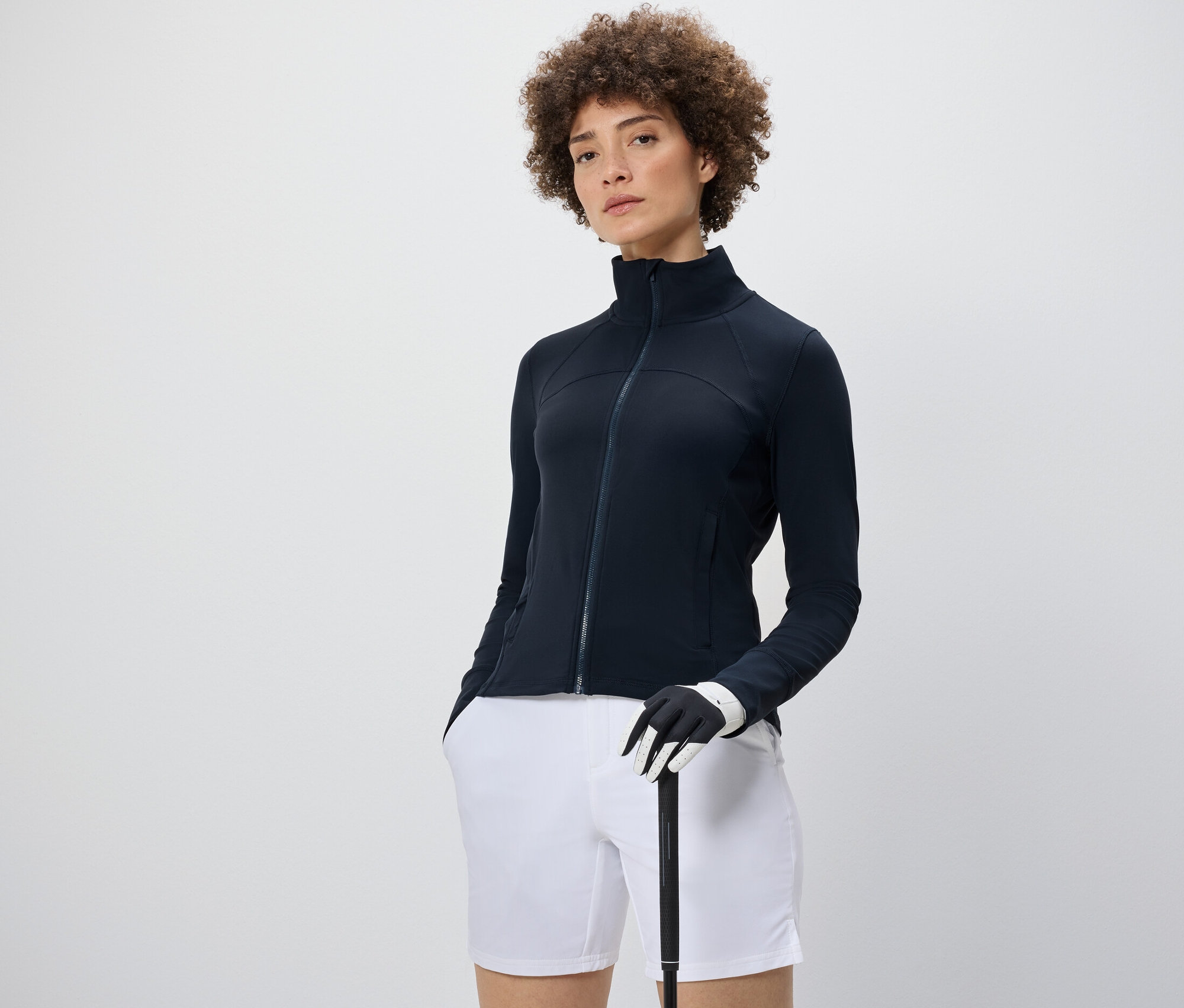 Nő visel egy Férfi funkcionális, kapucnis felső, krém/sötétkék színű felsőt, fehér rövidnadrágot és egy golfütőt tart.