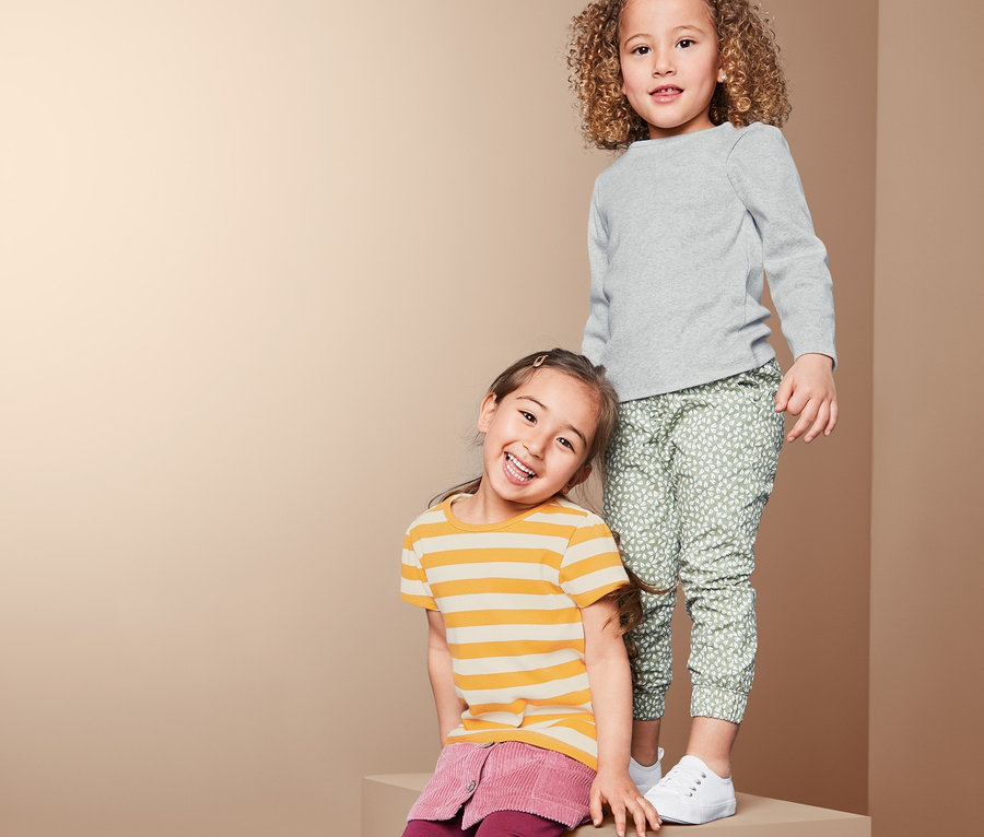 Két lány pózol egy tömbön. Az egyiken szürke bordázott felső, a másikon sárga csíkos póló és bordó szoknya van.