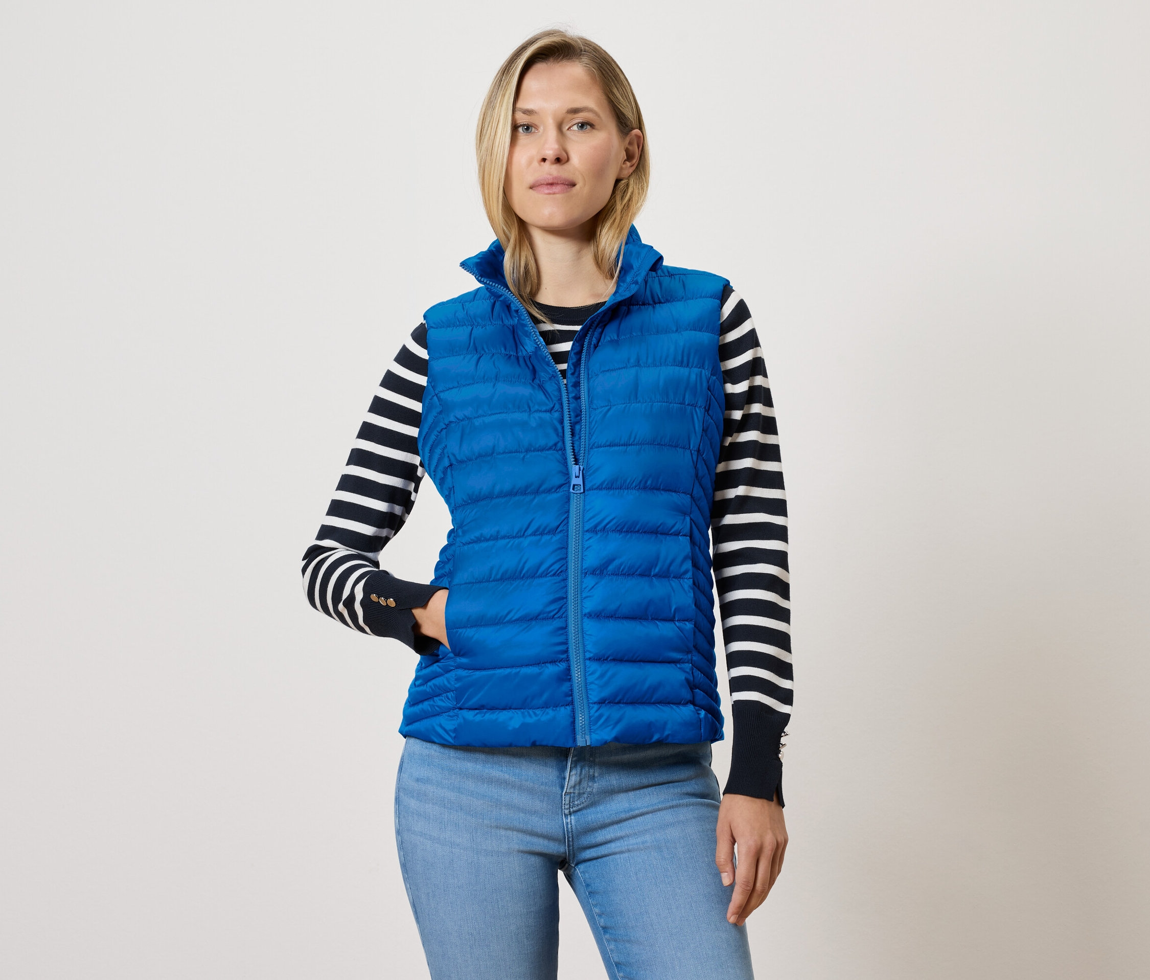 Nő kék, cipzáras, steppelt mellényt, fekete-fehér csíkos pulóvert és kék farmert visel.