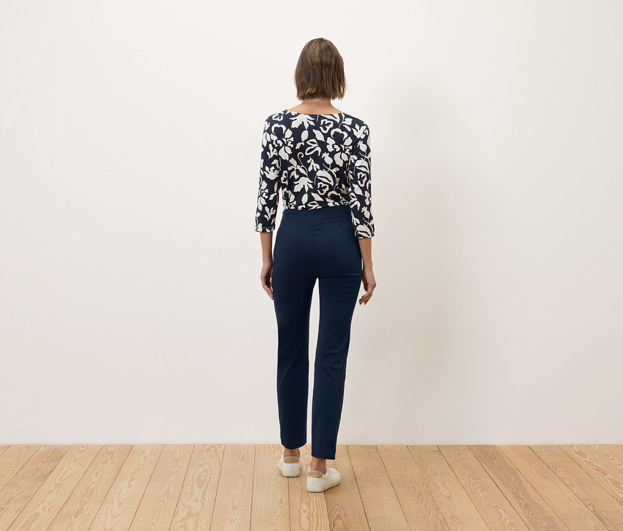 Egy barna hajú nő áll háttal a kamerának, tengerészkék nadrágot és kék-fehér virágmintás felsőt visel.