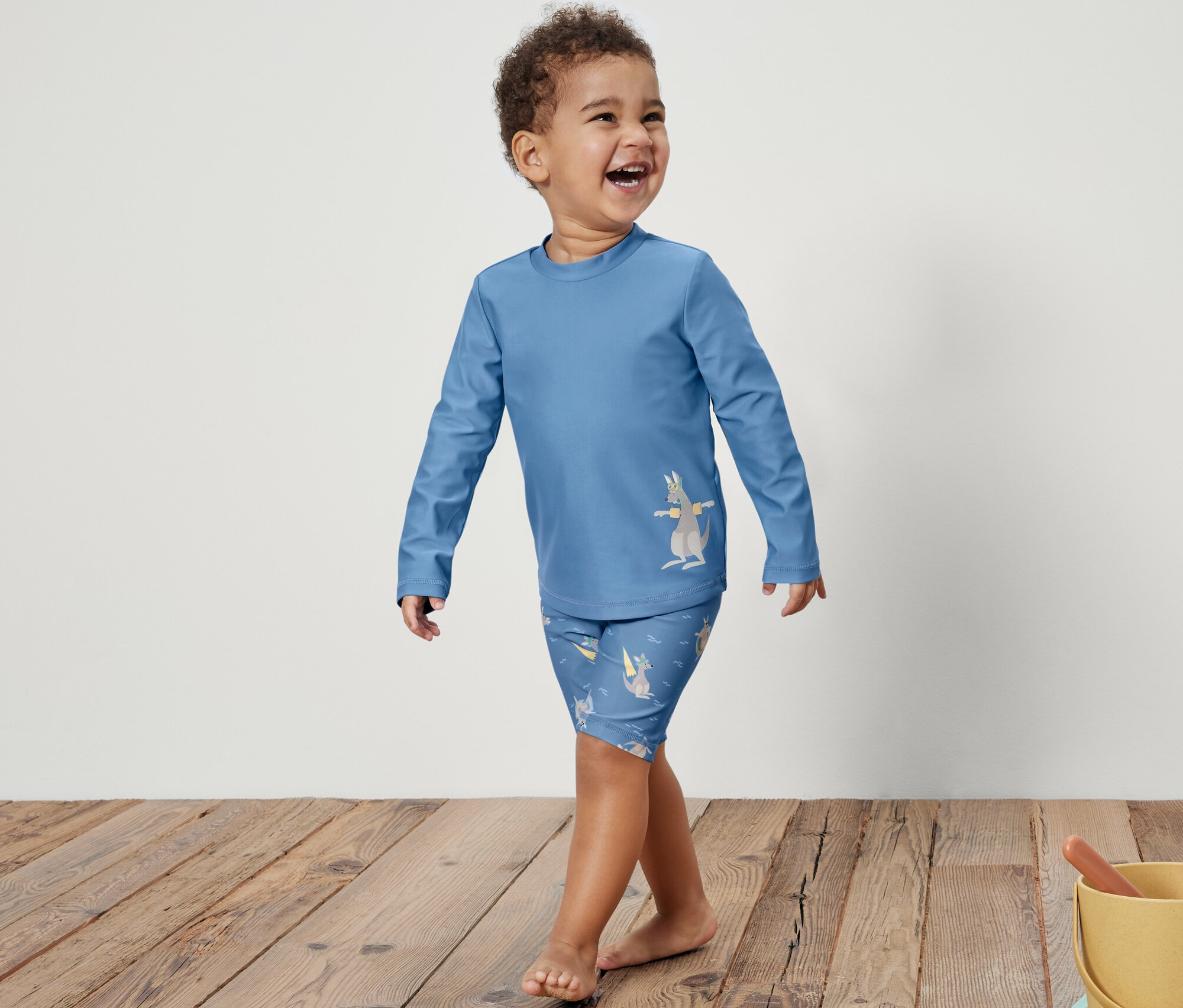 Egy mosolygó kisfiú kék, 80-as fényvédő faktorú, kenguru mintás gyerek együttesben sétál a fapadlón. Jobbra egy sárga vödör homokozó játékokkal.