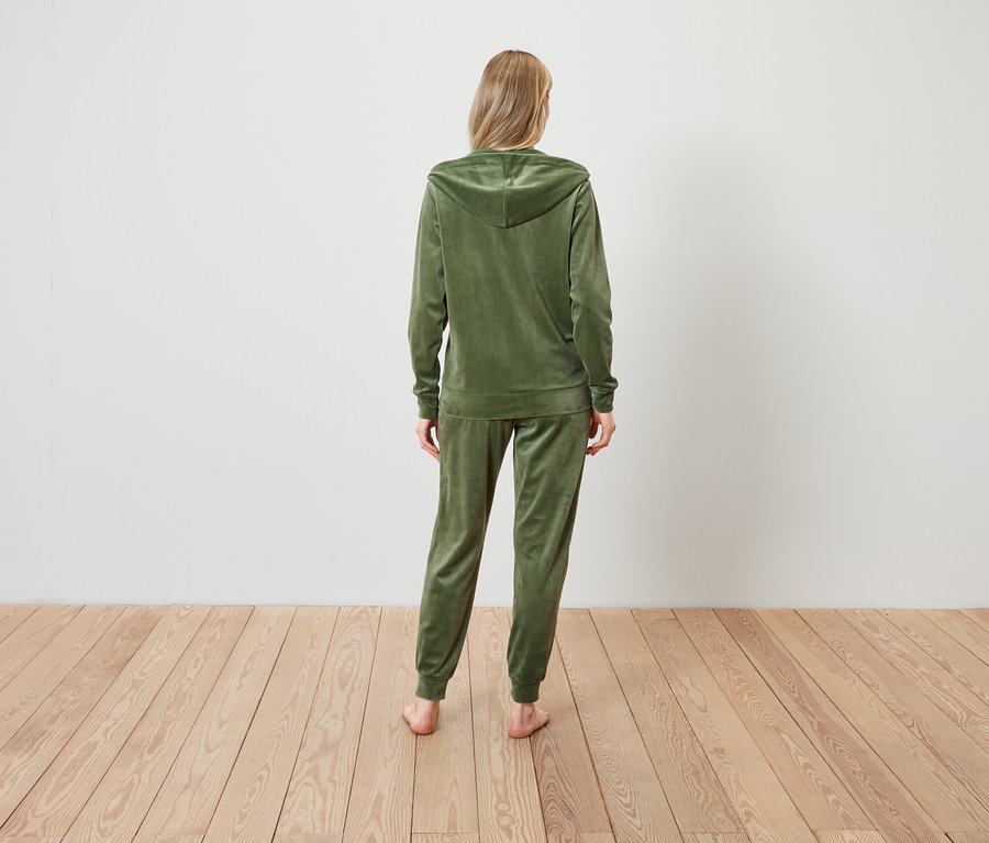 Egy nő áll háttal a kamerának, olívazöld színű velúr tréningruhát visel.
