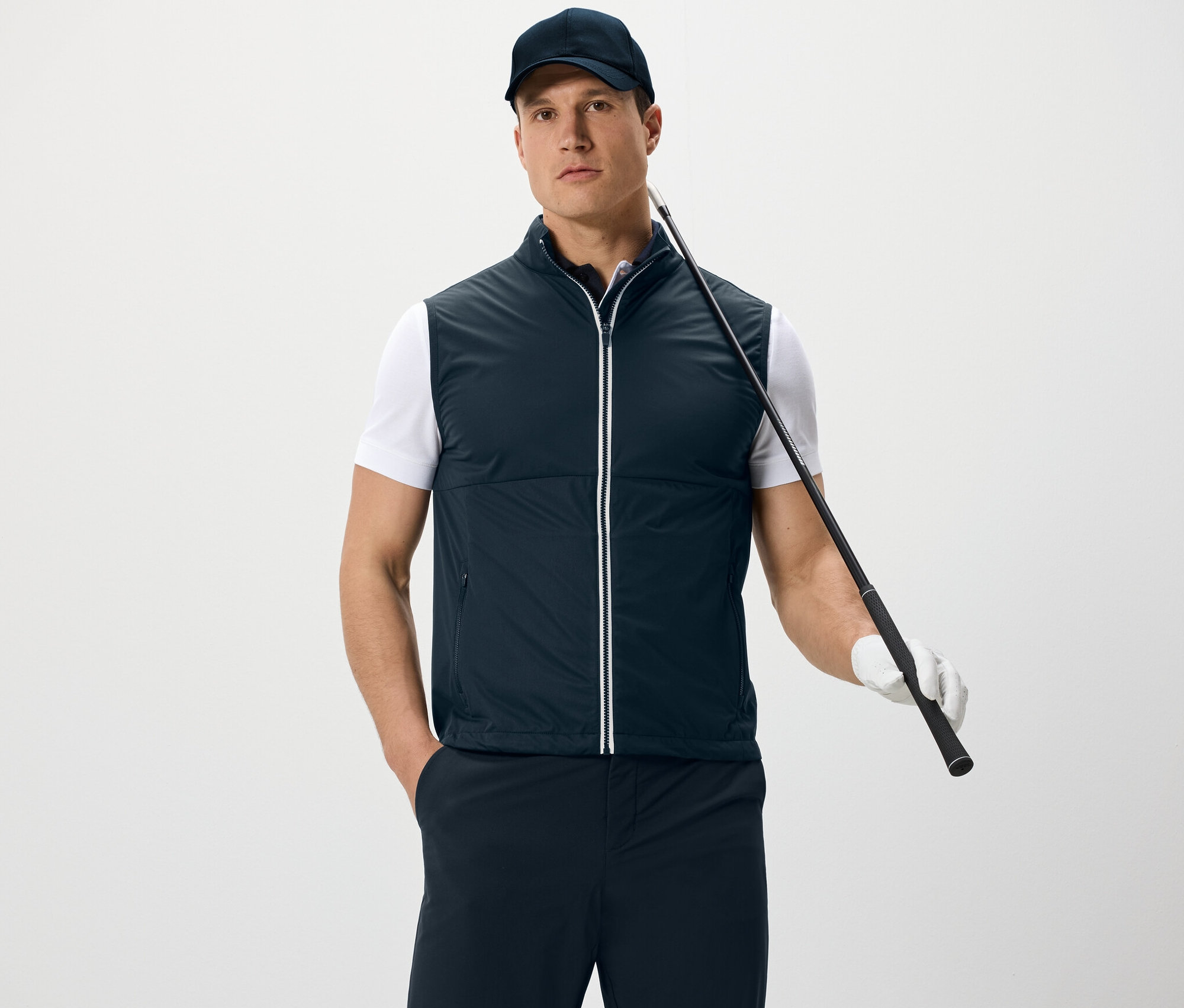 Férfi softshell mellényt és baseballsapkát visel, golfütőt tart, és fehér háttér előtt áll.