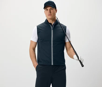 Férfi softshell mellényt és baseballsapkát visel, golfütőt tart, és fehér háttér előtt áll.