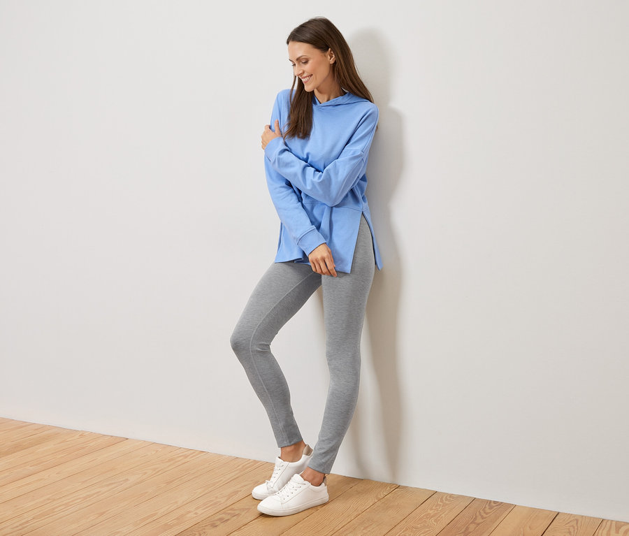 Nő a falnak támaszkodik, világoskék, oldalt sliccelt kapucnis pulóvert, szürke leggingset és fehér tornacipőt visel.